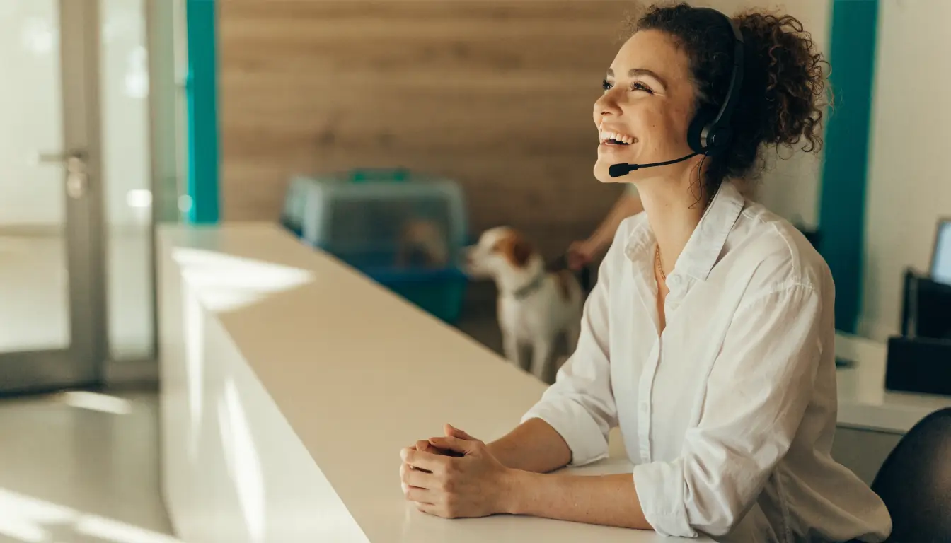 Illustrative stock photo: a smiling receptionist wearing a headset at a clinic front desk, a dog waiting in the background.
